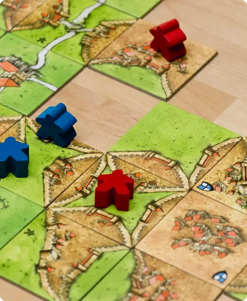 Een opstelling van het bordspel Carcassonne met groene landschapstegels en strategisch geplaatste rode en blauwe pionnen. Het tafereel straalt strategisch spel uit