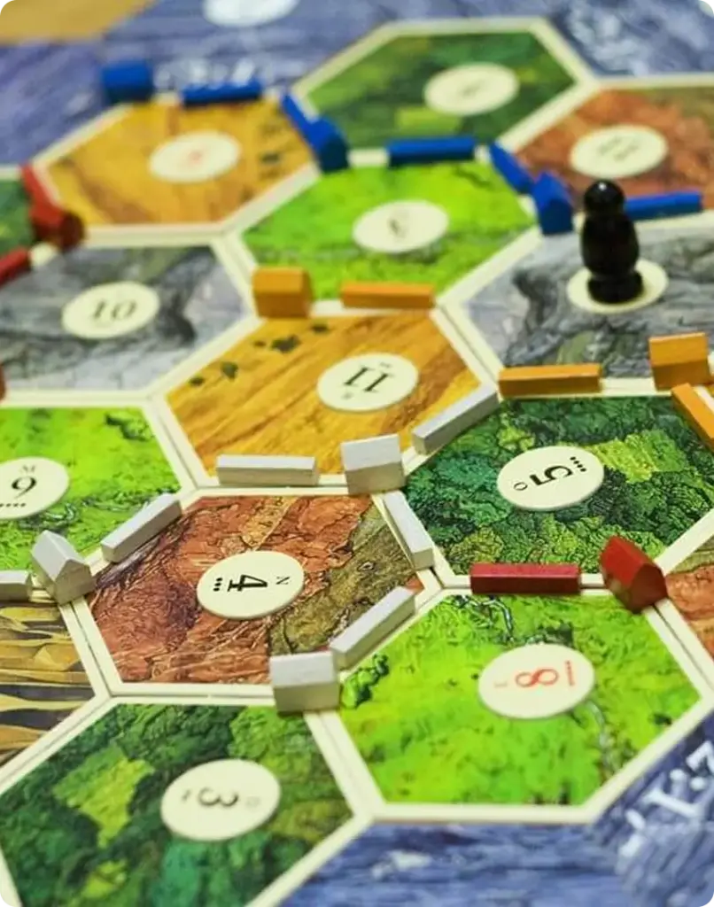 Een bordspel van „De Kolonisten van Catan“ in volle gang, met zeshoekige tegels met cijfers en gekleurde wegen. In het midden staat een zwart speelstuk dat de voortgang aangeeft